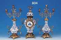 VASENUHR MIT ZWEI BEISTELLERN, WOHL FLANDERN, UM 1920/30, KERAMIK MIT BLAUER CAMAIEU-MA- LEREI, MESSINGMONTUREN, GLASDECKEL, EMAILZIF- FERBLATT MIT ...