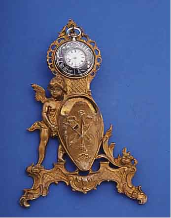 UHRENSTäNDER DARSTELLUNG AMOR, PANTOFFEL HAL- TEND UMGEB. M. ÖSE RüCKS. ALS WANDANH., MES-SING VERG., BER., UM 1890, H. CA. 22 CM, ...