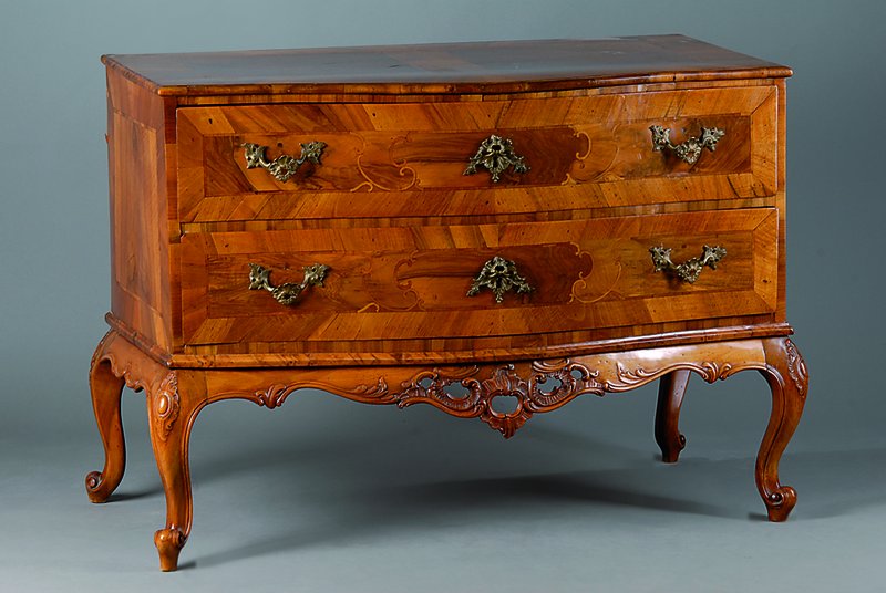 Kommode, um 1760/70, Nußbaumfurnier, 2- schübig, Front geschweift, Zarge mit reicher Schnitzerei in Durchbrucharbeit, Intarsien in Volutenform, ...