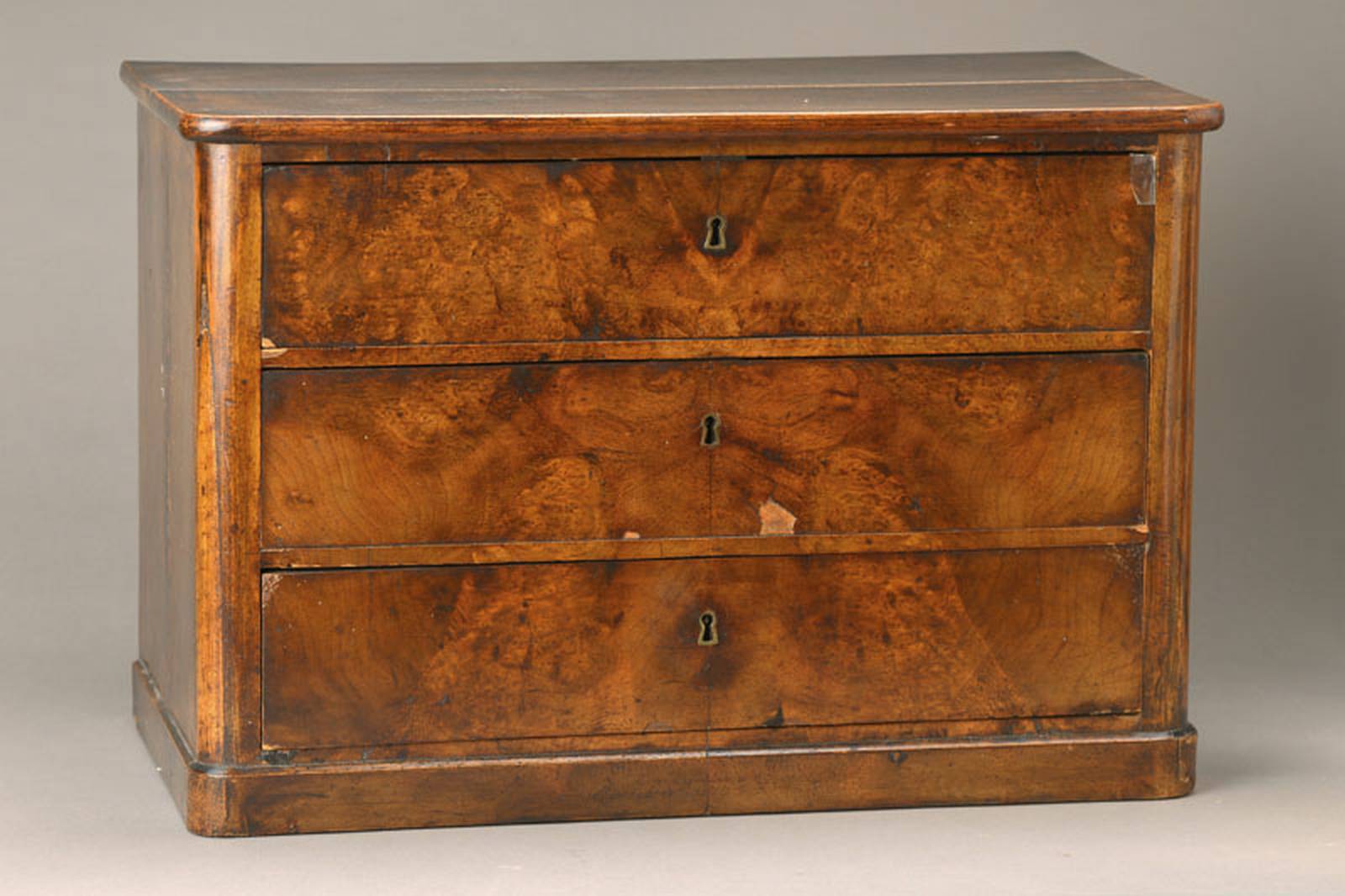 Miniaturkommode, südwestdeutsch, wohl Baden, um 1830/40, Nussbaumfurnier, Weichholzkorpus,abgerundete Ecken, 3-schübig, Furniersch., schwundrissig, ...