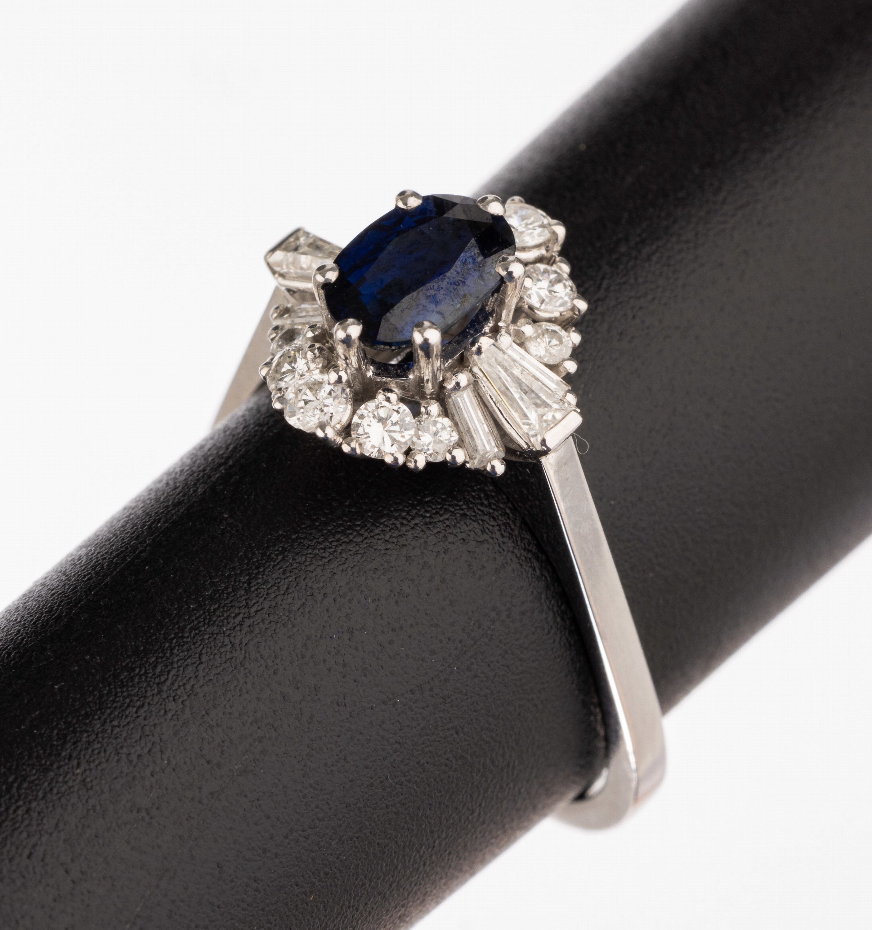 18 kt Gold Saphir-Diamant-Ring, WG 750/000, mittig ovalfacett. Saphir ca. 0.90 ct, umgeben von 10 Brillanten, 2 Trapeze und 4Baguettes zus. ca. 0.65 ...