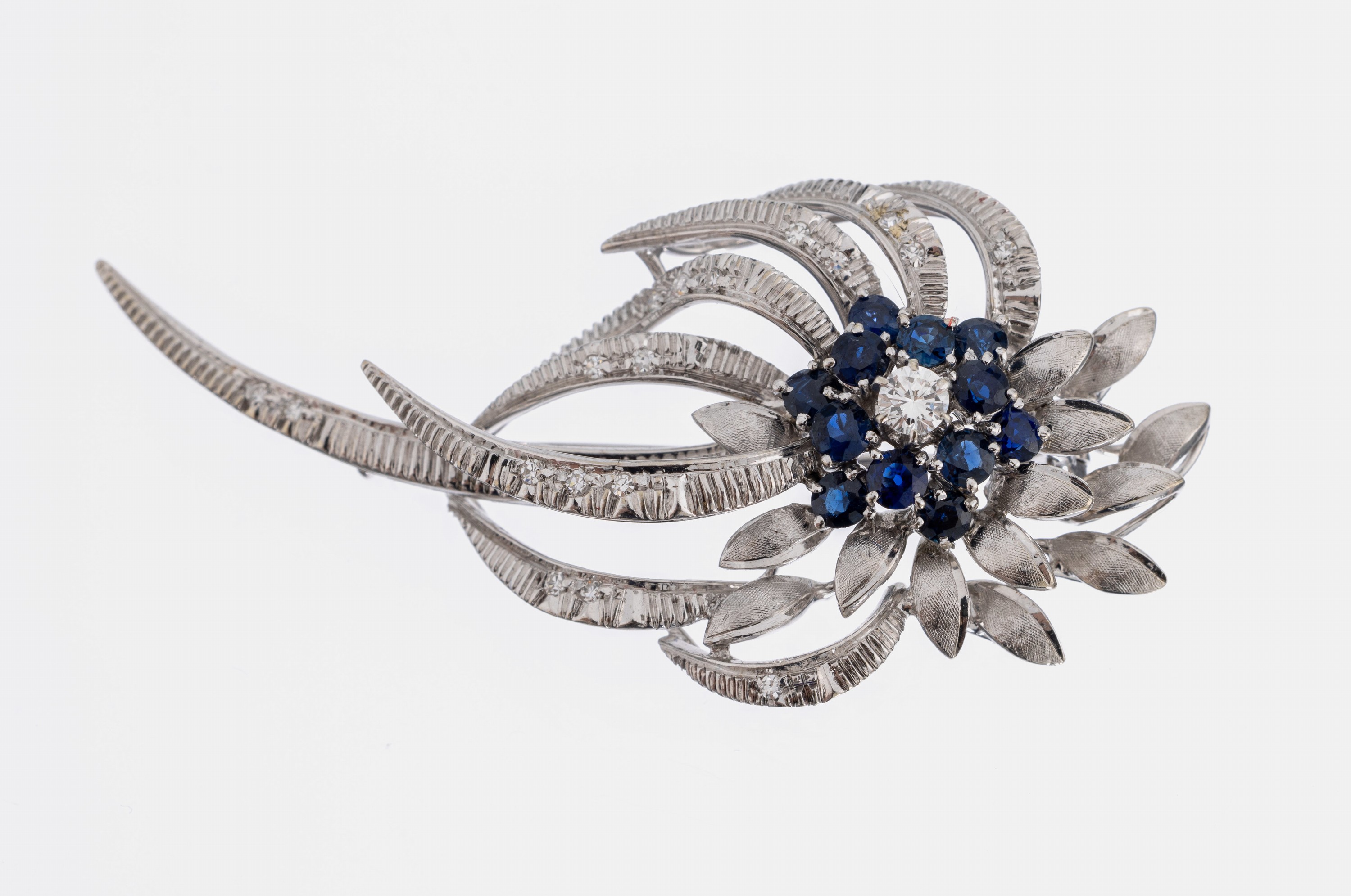 18 kt Gold Saphir Brillant Brosche, WG 750/000, Blütenform, teilmatt. und struk- turiert, 12 rundfacett. Saphire zus. ca. 1.0 ct, 1 Brillant ca. ...