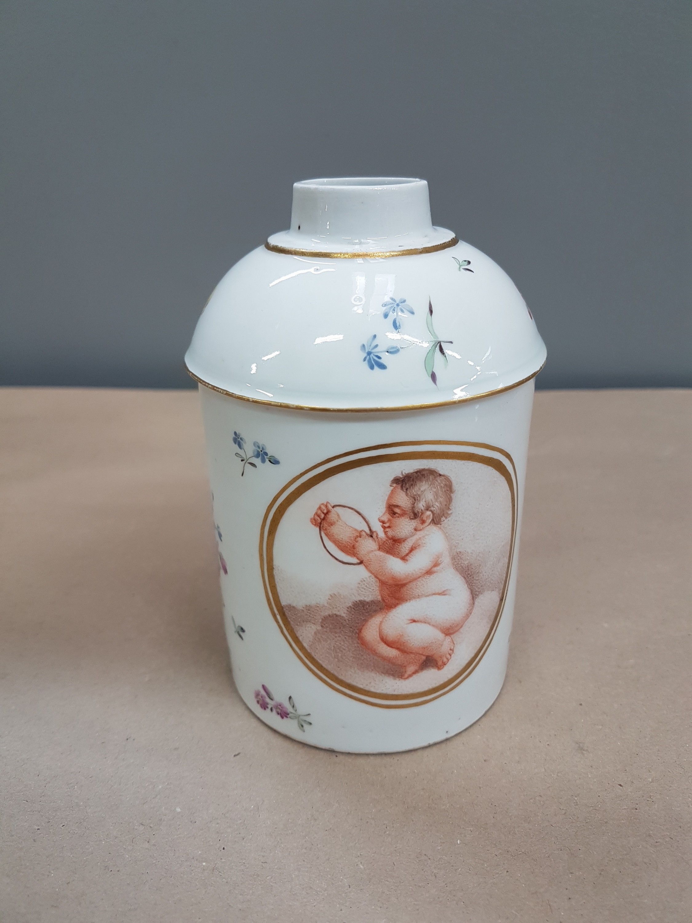Detail c: Große Teedose Frankenthal, dat. 1779, CT-Marke, Porzellan, im Oval, sitzender Putto mit rotem Mantel, gestreute Blümchen, ohne Deckel, H. ca. 13 cm