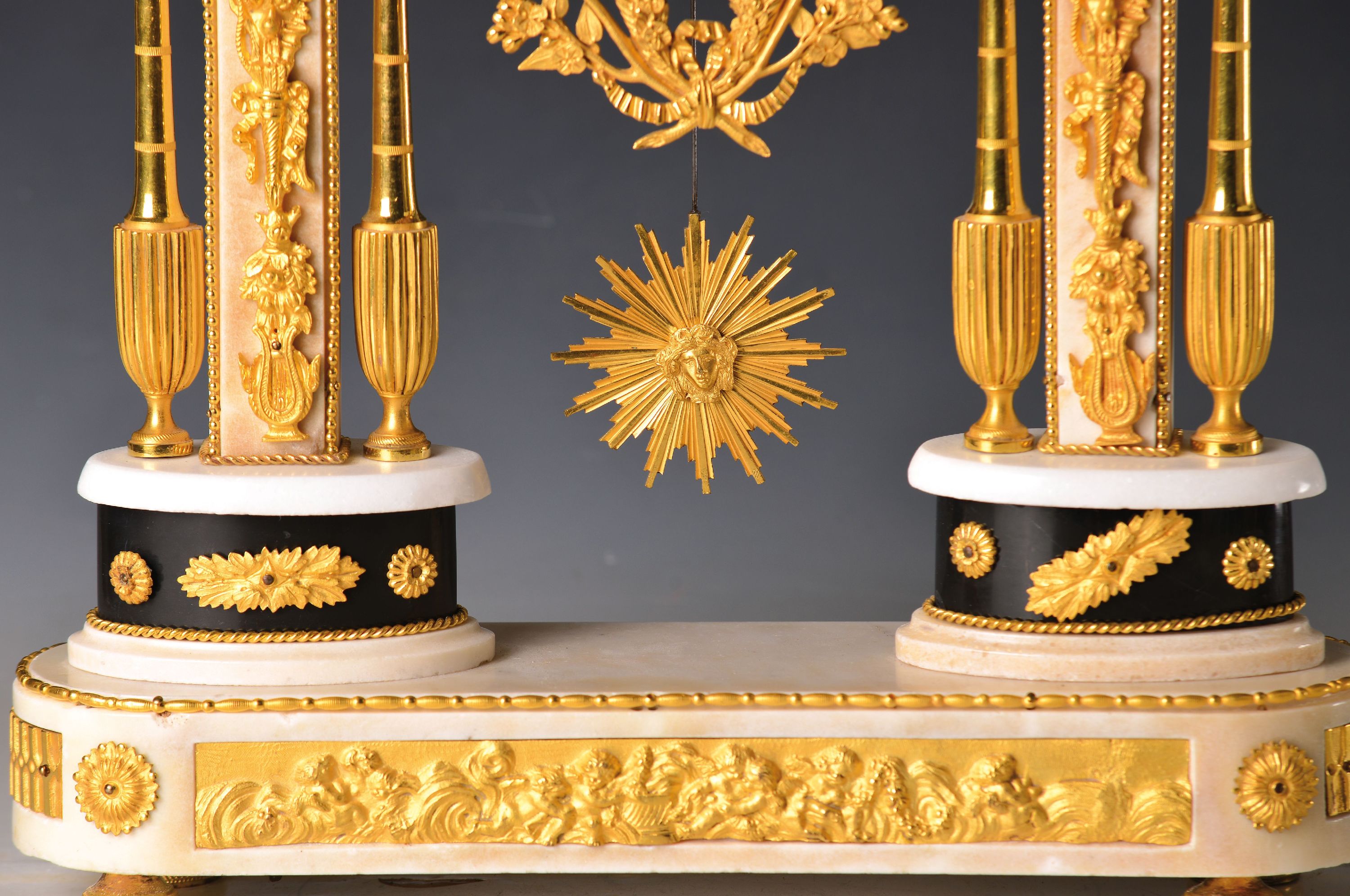 Detail b: Portaluhr, Dessille a Paris, um 1790, feuervergoldete Appliken auf weißem Marmor, Sockel mit reliefierten Putti, darauf zwei ovale Plinthen mit ...