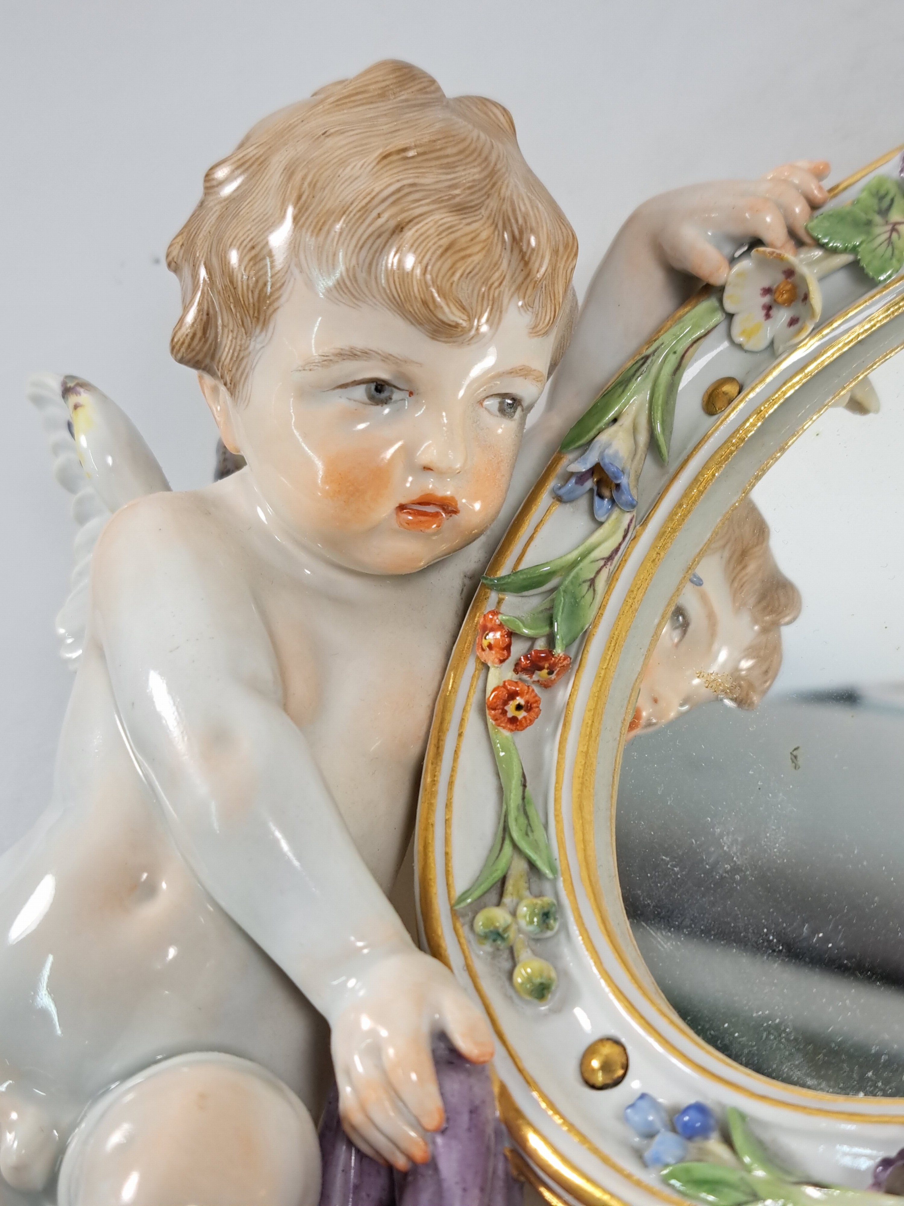 Detail c: Wandspiegel mit Amorette, Meissen um 1900, Porzellan, Amorette mit lilafarbener Schärpe hält eine ovalen Spiegel, applizierte Blüten, polychrom ...
