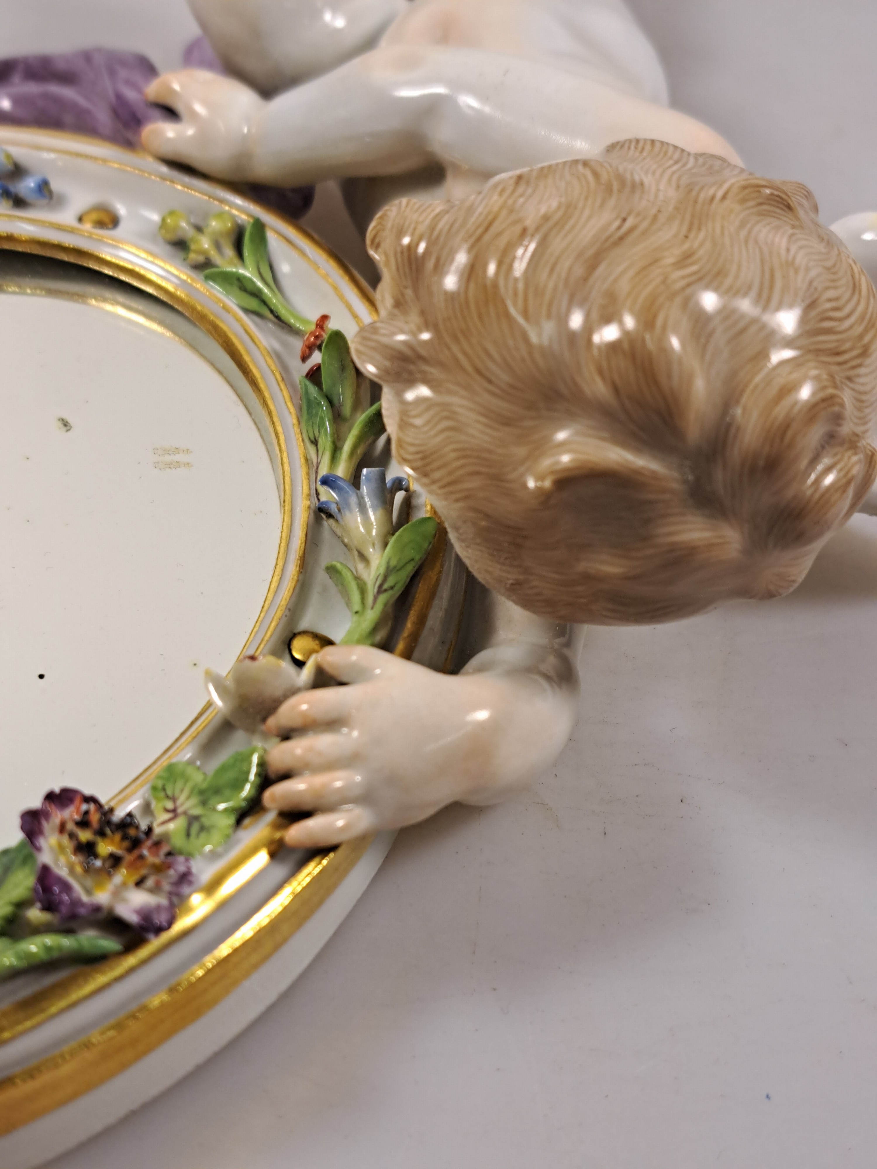Detail i: Wandspiegel mit Amorette, Meissen um 1900, Porzellan, Amorette mit lilafarbener Schärpe hält eine ovalen Spiegel, applizierte Blüten, polychrom ...
