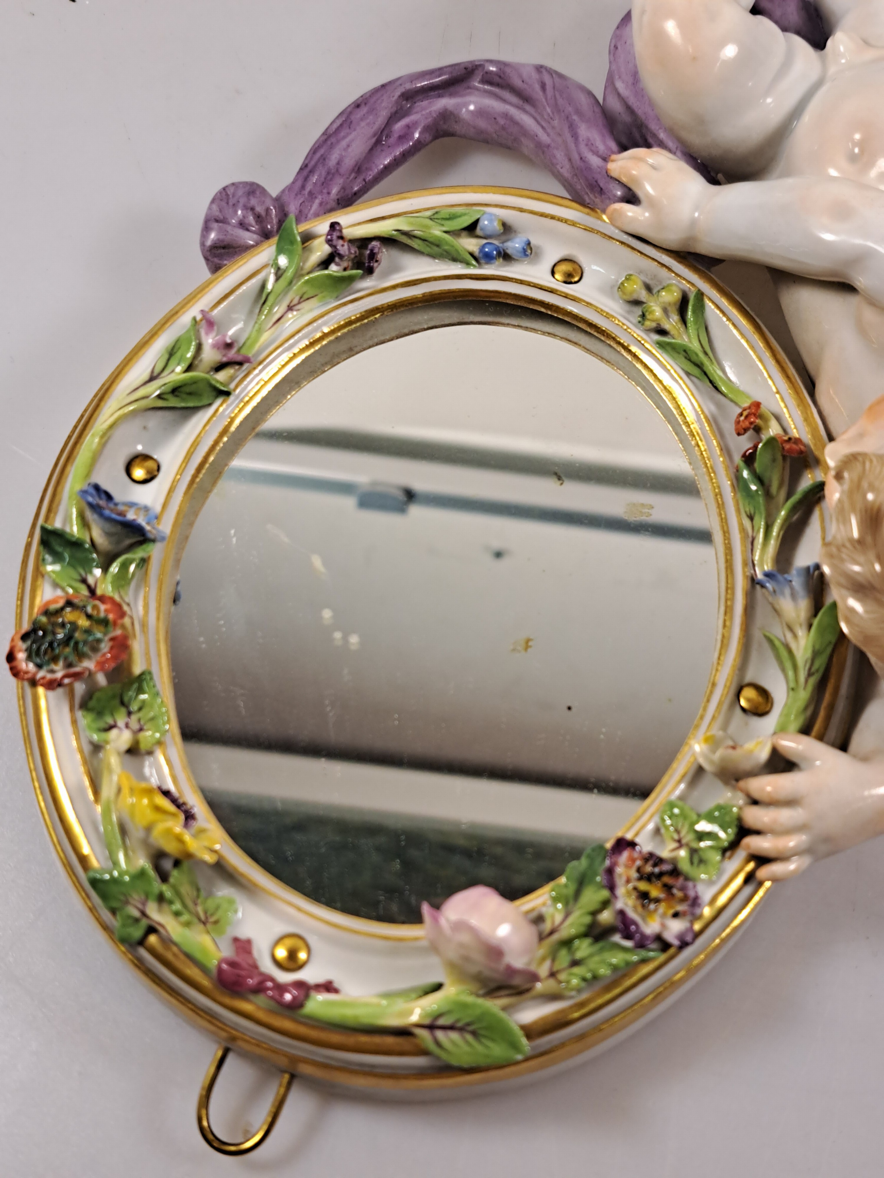 Detail j: Wandspiegel mit Amorette, Meissen um 1900, Porzellan, Amorette mit lilafarbener Schärpe hält eine ovalen Spiegel, applizierte Blüten, polychrom ...