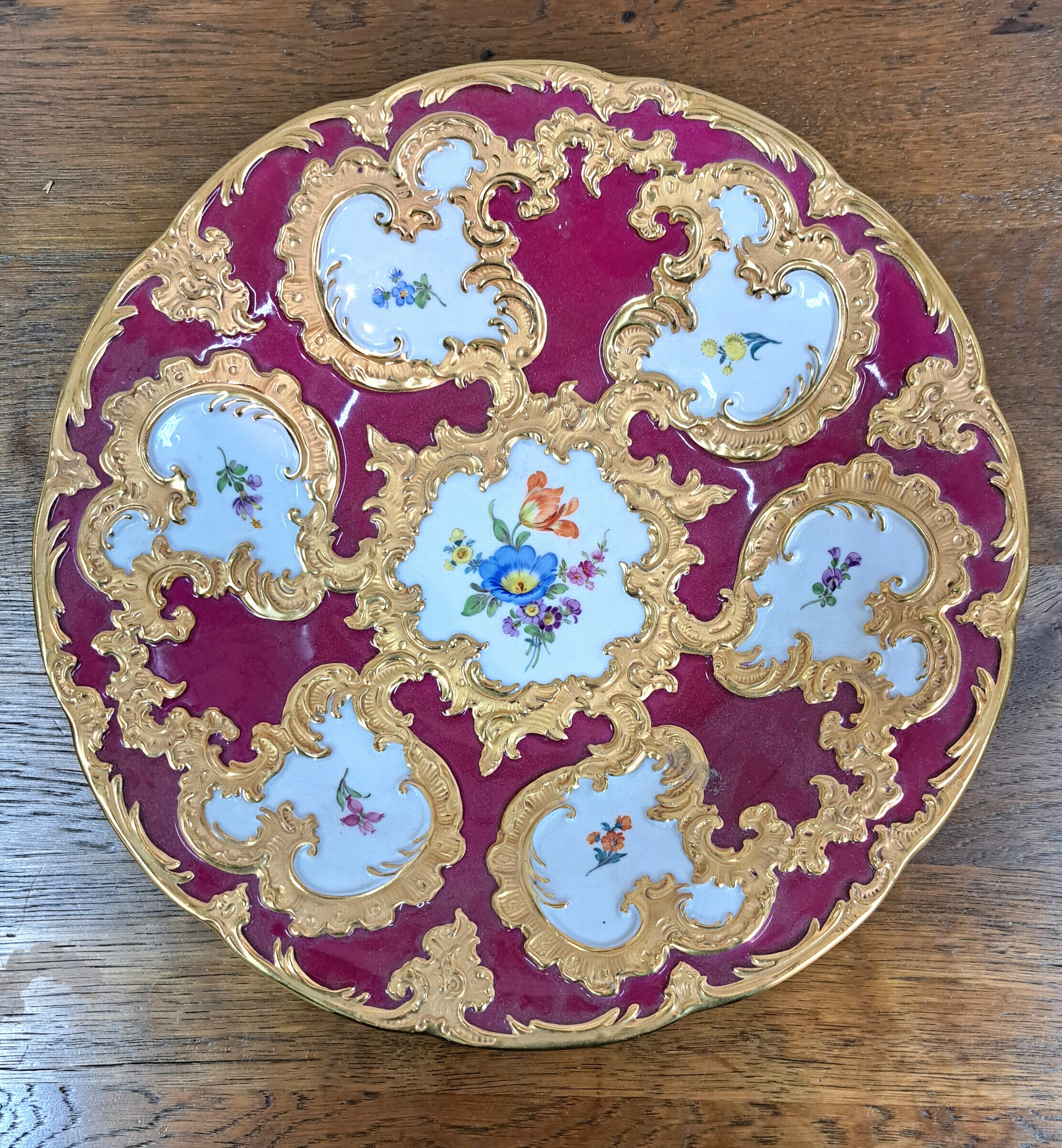 Detail a: Prunkteller, Meissen, 20. Jh., 1. Wahl, partiell dunkelrot glasiert, reich reliefiert und matt- und glanzvergoldet, bunte Blumen, D. ca. 28cm