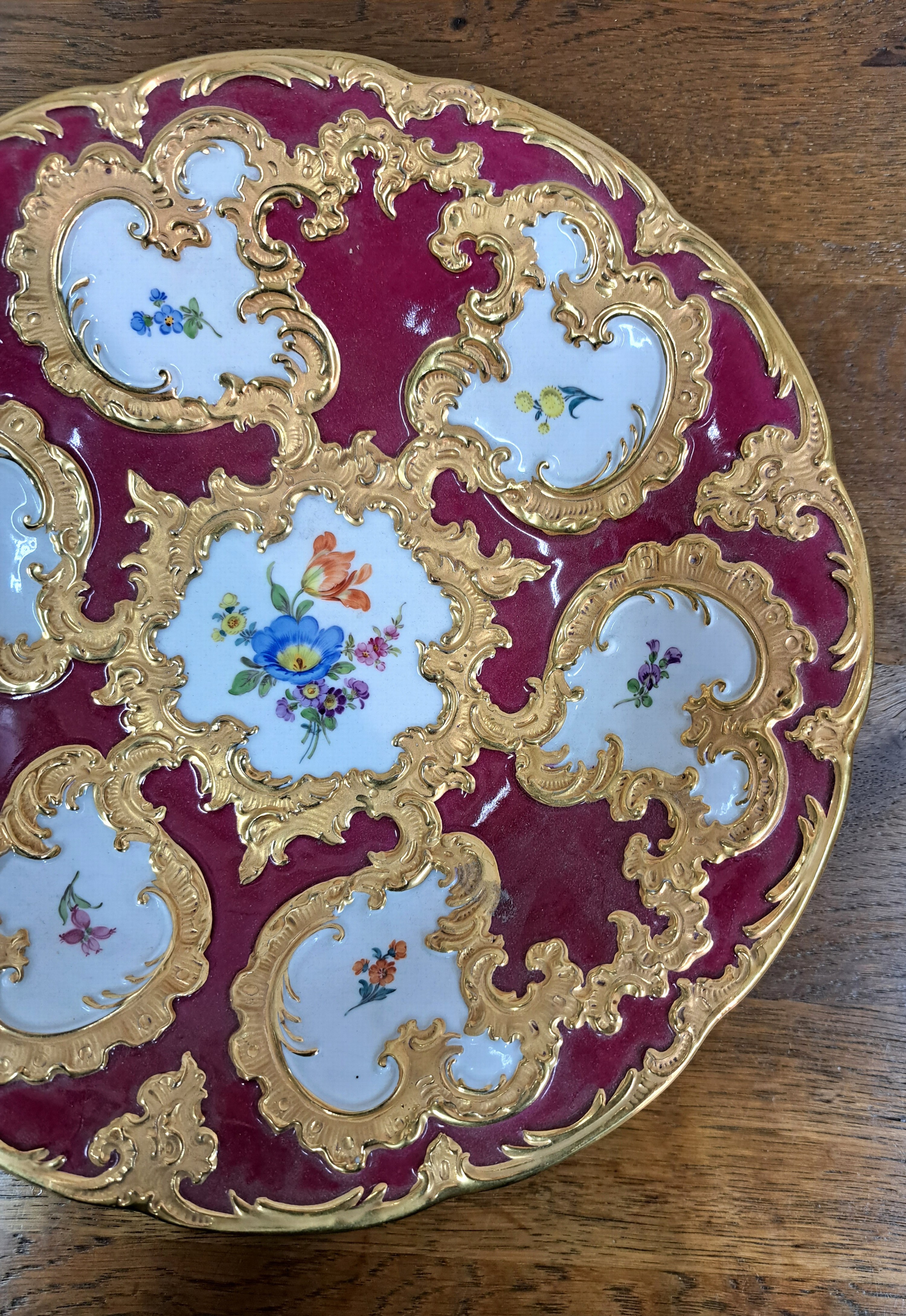 Detail b: Prunkteller, Meissen, 20. Jh., 1. Wahl, partiell dunkelrot glasiert, reich reliefiert und matt- und glanzvergoldet, bunte Blumen, D. ca. 28cm
