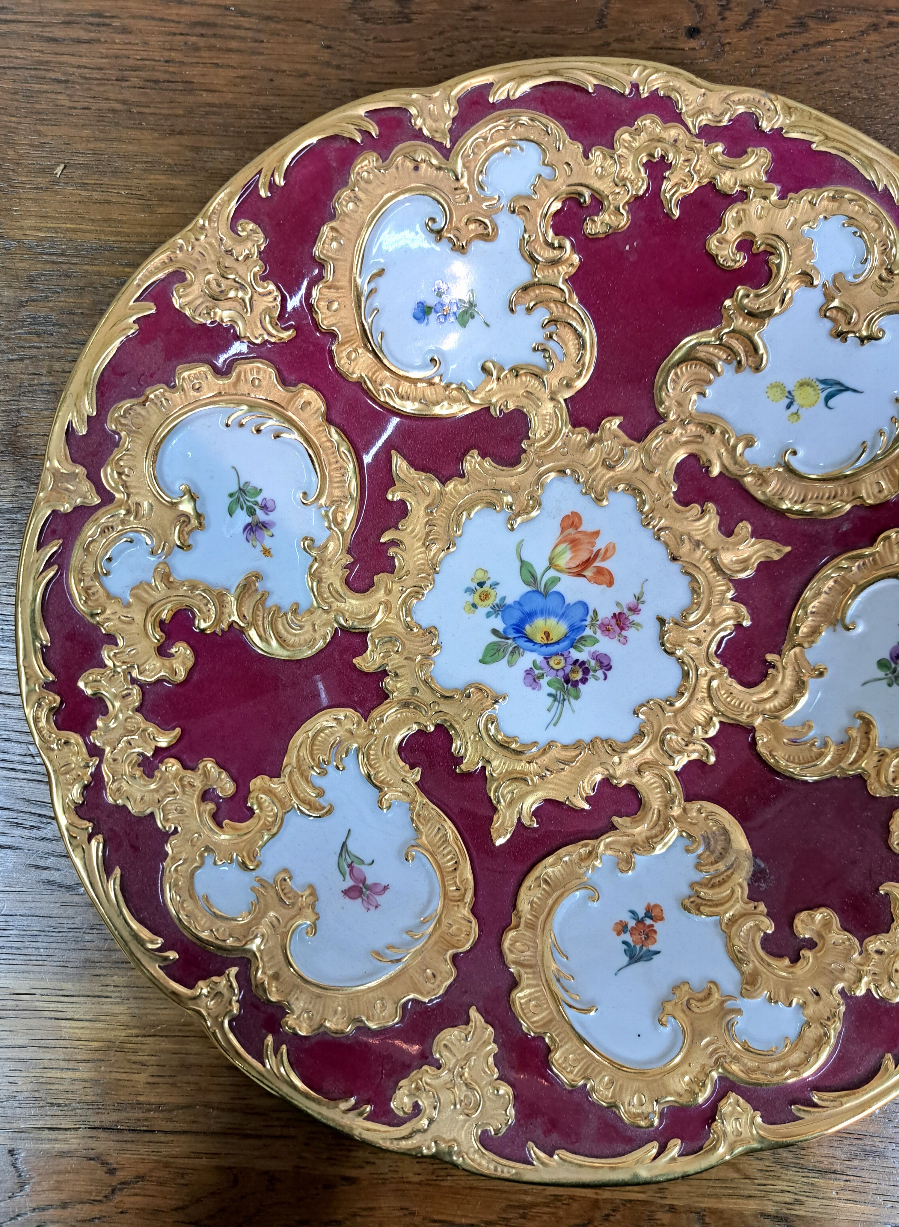 Detail c: Prunkteller, Meissen, 20. Jh., 1. Wahl, partiell dunkelrot glasiert, reich reliefiert und matt- und glanzvergoldet, bunte Blumen, D. ca. 28cm